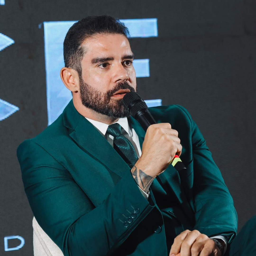 Homem de barba, terno verde e gravata, sentado em poltrona branca, fala ao microfone diante de um telão