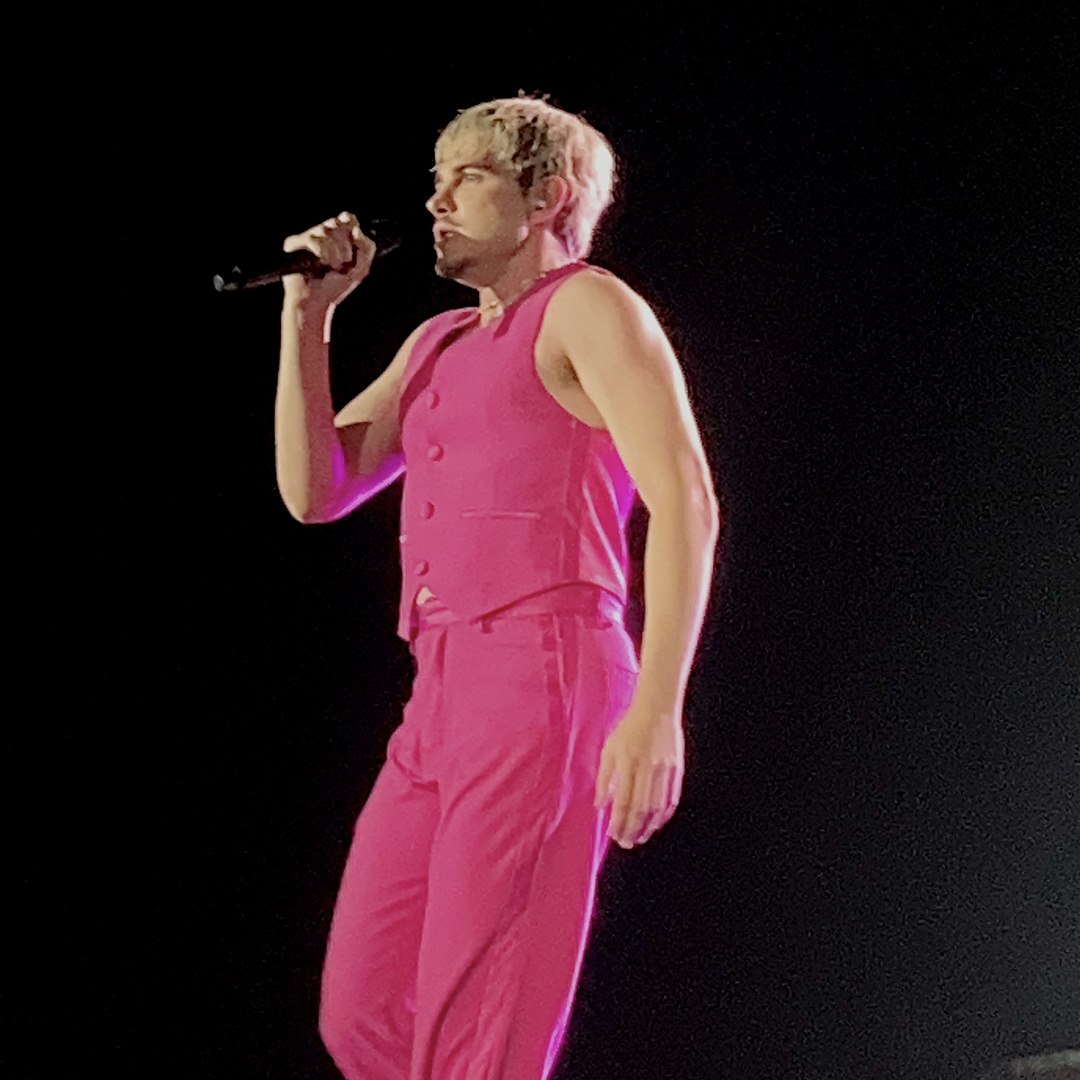 Jão canta ao microfone no palco, vestindo colete e calça cor-de-rosa
