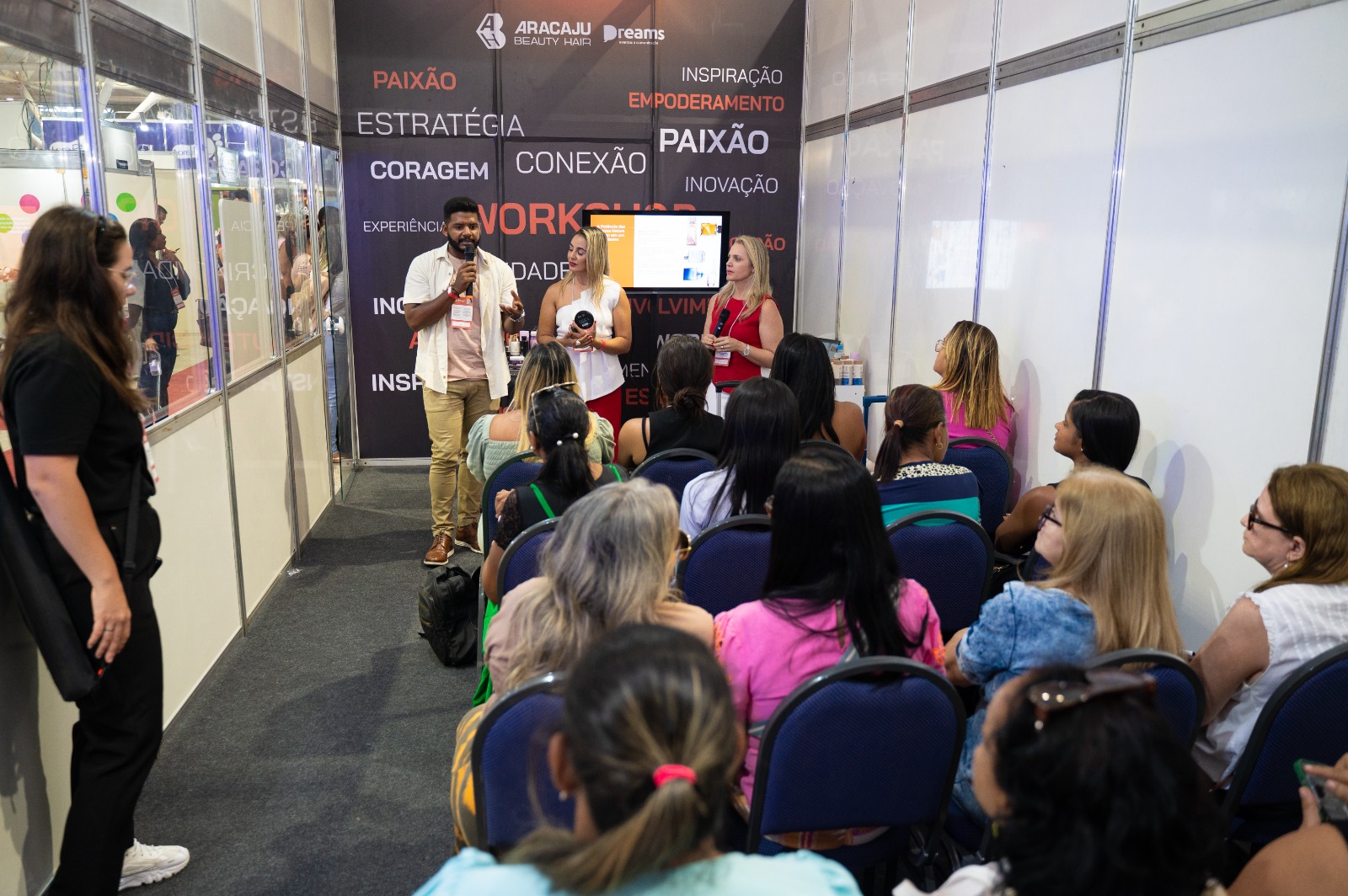 Três palestrantes falam ao microfone para plateia sentada em sala de workshop com painel da Aracaju Beauty Hair e da Dreams