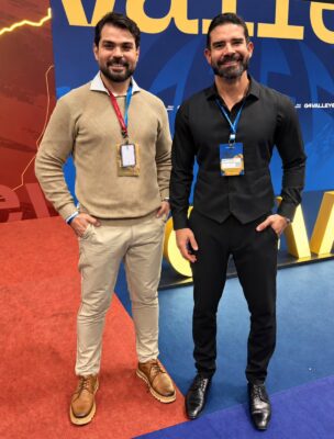 Dois homens de barba sorriem lado a lado com crachás do evento, um de suéter bege e calça clara e o outro todo de preto, diante do painel azul e amarelo do G4 Valley