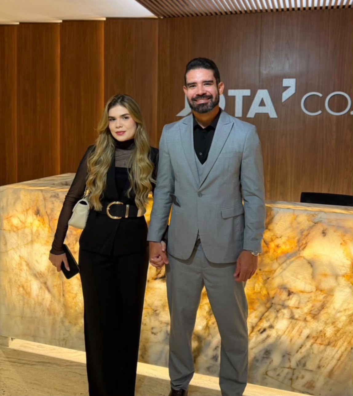 Casal de mãos dadas, ela de roupa preta e cabelos longos e ele de terno cinza, diante de um balcão de mármore iluminado com a marca do Jota Jota Podcast