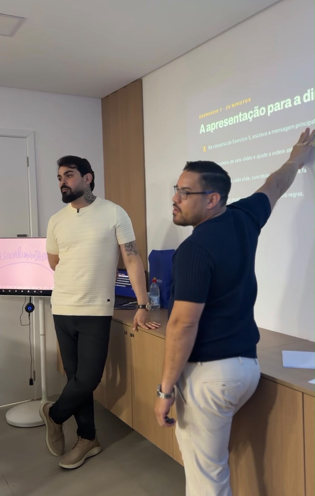 Dois homens conduzem uma aula em sala com projeção de exercício na parede e uma tela com o nome Descomplicando IA ao lado