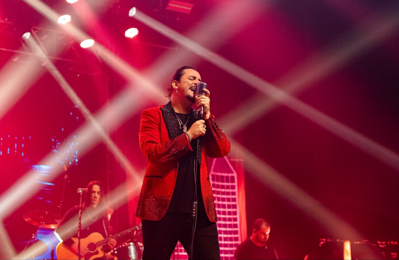 O cantor Dino Fonseca, de paletó vermelho brilhante, canta em um microfone retrô no palco, com luzes vermelhas e um violonista ao fundo