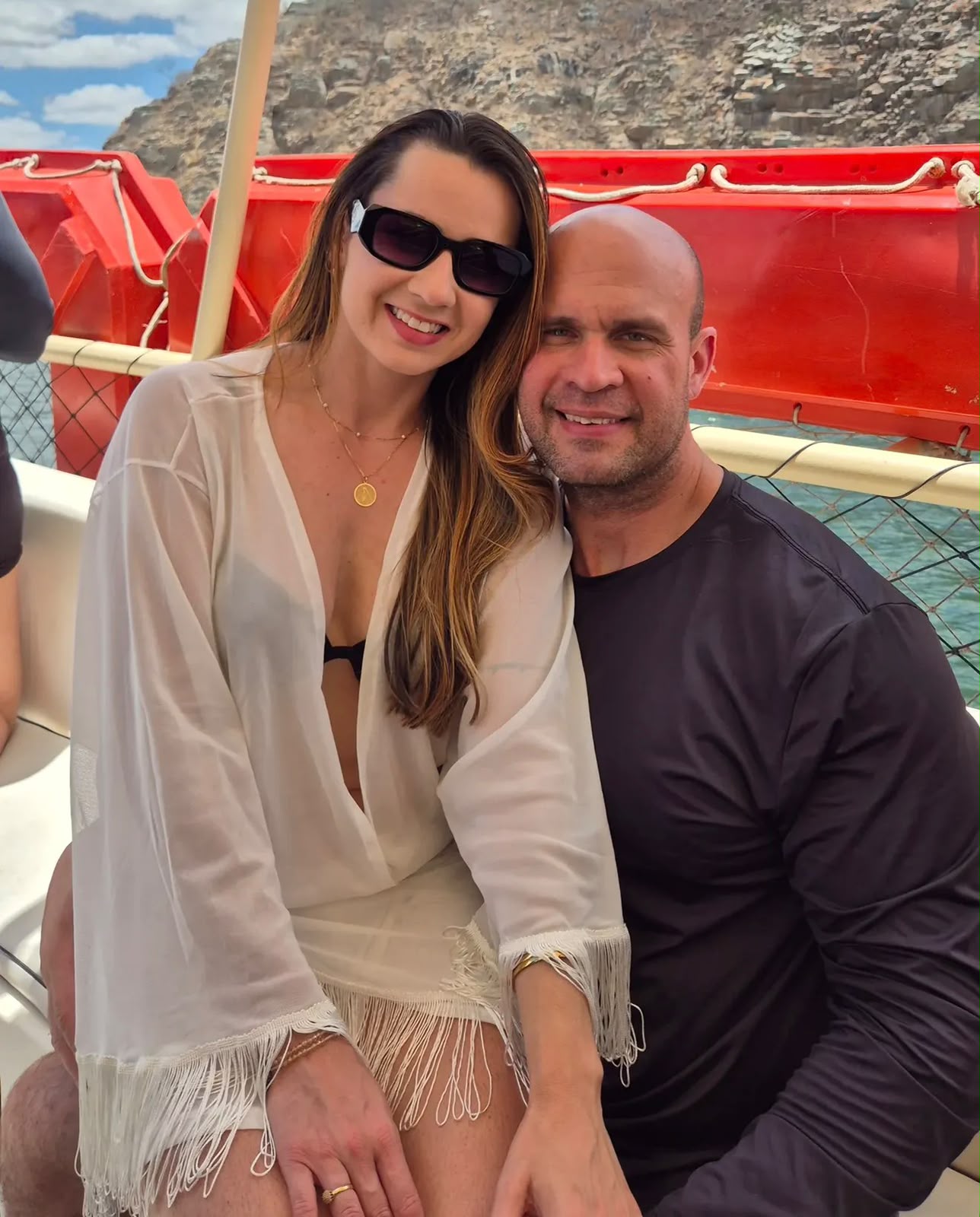Dr. Adelmo Alves, de camiseta escura, sorri abraçado à esposa Jhennefer Ramos, de óculos escuros e saída de praia branca, sentados em um barco com paredões de pedra ao fundo