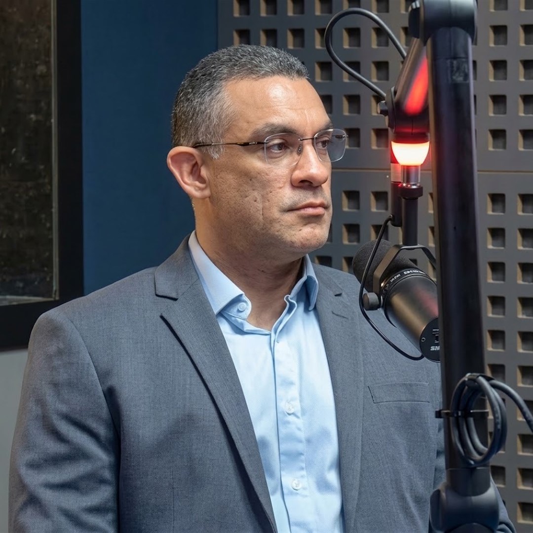 Homem de óculos, camisa azul-clara e paletó cinza em estúdio de rádio, ao lado de um microfone com luz vermelha acesa
