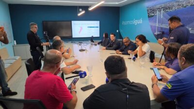 Pessoas reunidas em torno de uma mesa em sala da Iguá Sergipe, com microfones de imprensa e um telão exibindo mapa das áreas afetadas