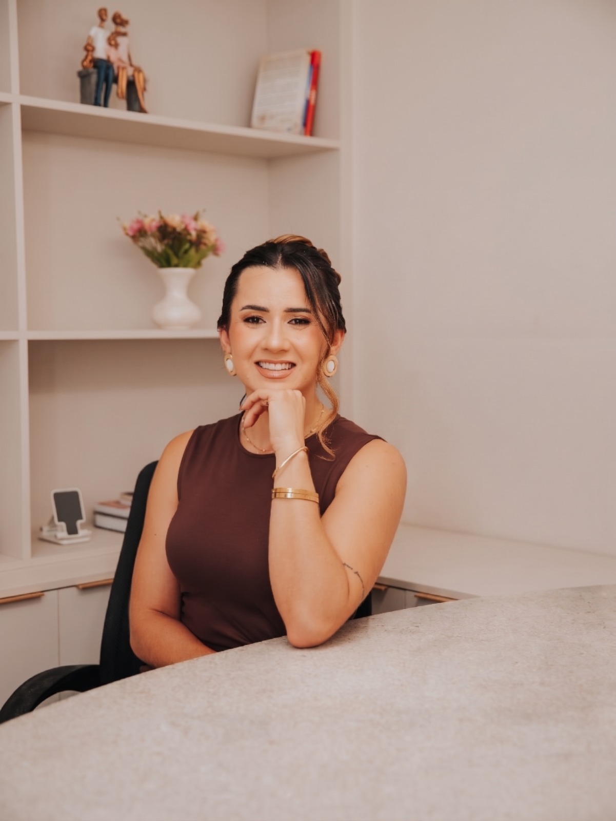 Jhenefer Ramos, de blusa marrom sem mangas, sorri com a mão apoiada no queixo, sentada a uma mesa clara em um escritório com estante e flores ao fundo