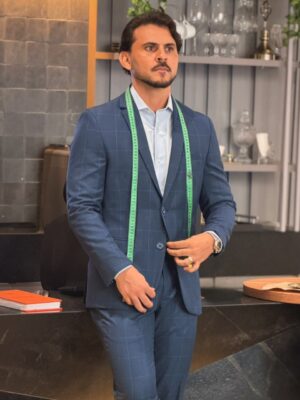 Leandro Andrade de terno azul xadrez e camisa clara, com fita métrica verde sobre os ombros, em ambiente de ateliê