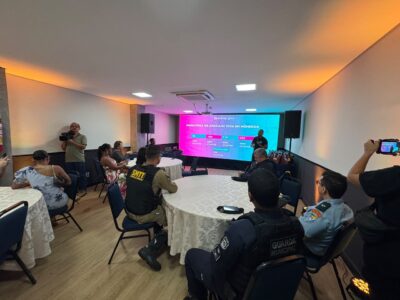 Coletiva de imprensa em salão com mesas redondas, cinegrafista e agentes de trânsito e da Guarda Municipal, diante de telão com os números da Maratona de Aracaju 2026