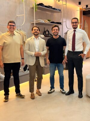 Quatro homens sorrindo lado a lado dentro de uma loja de moda masculina, com prateleiras de roupas e sapatos ao fundo