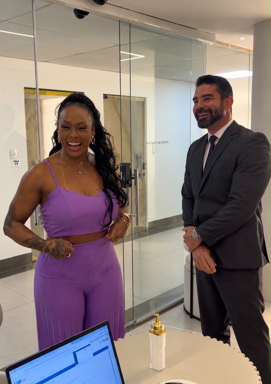 Rosiane Pinheiro, de conjunto lilás, gargalha ao lado do Dr. Marlon Batista, de terno escuro e barba, em uma recepção