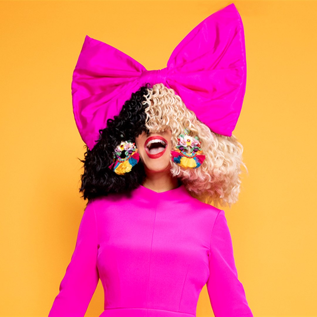 “Chandelier” e “Titanium” entram em venda milionária do catálogo de Sia 1 Sia com peruca metade preta e metade loira, laço rosa gigante na cabeça e vestido pink, em fundo laranja