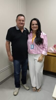 Homem de óculos e camisa polo preta posa ao lado de uma mulher de cabelos longos, blusa rosa listrada e calça branca, em uma sala